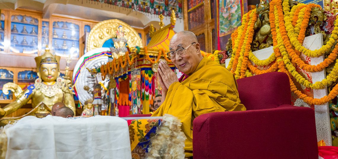 Sua Santidade o 14º Dalai Lama sorrindo, com as mãos em saudação, diante de um altar enfeitado com guirlandas de flores